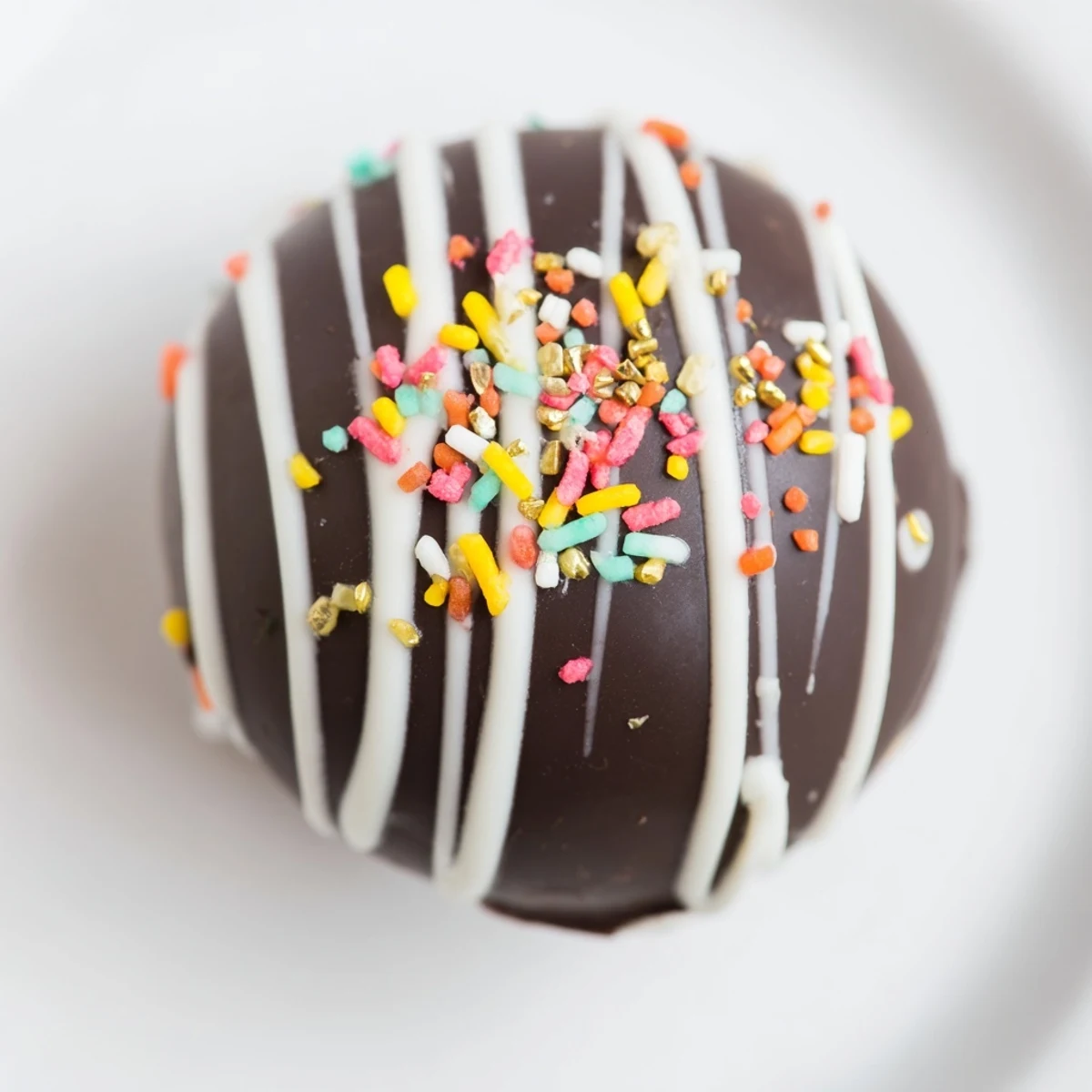 Glazed Hot Chocolate Bombs decorated with white chocolate drizzle and colorful sprinkles sit on a rustic wooden surface.